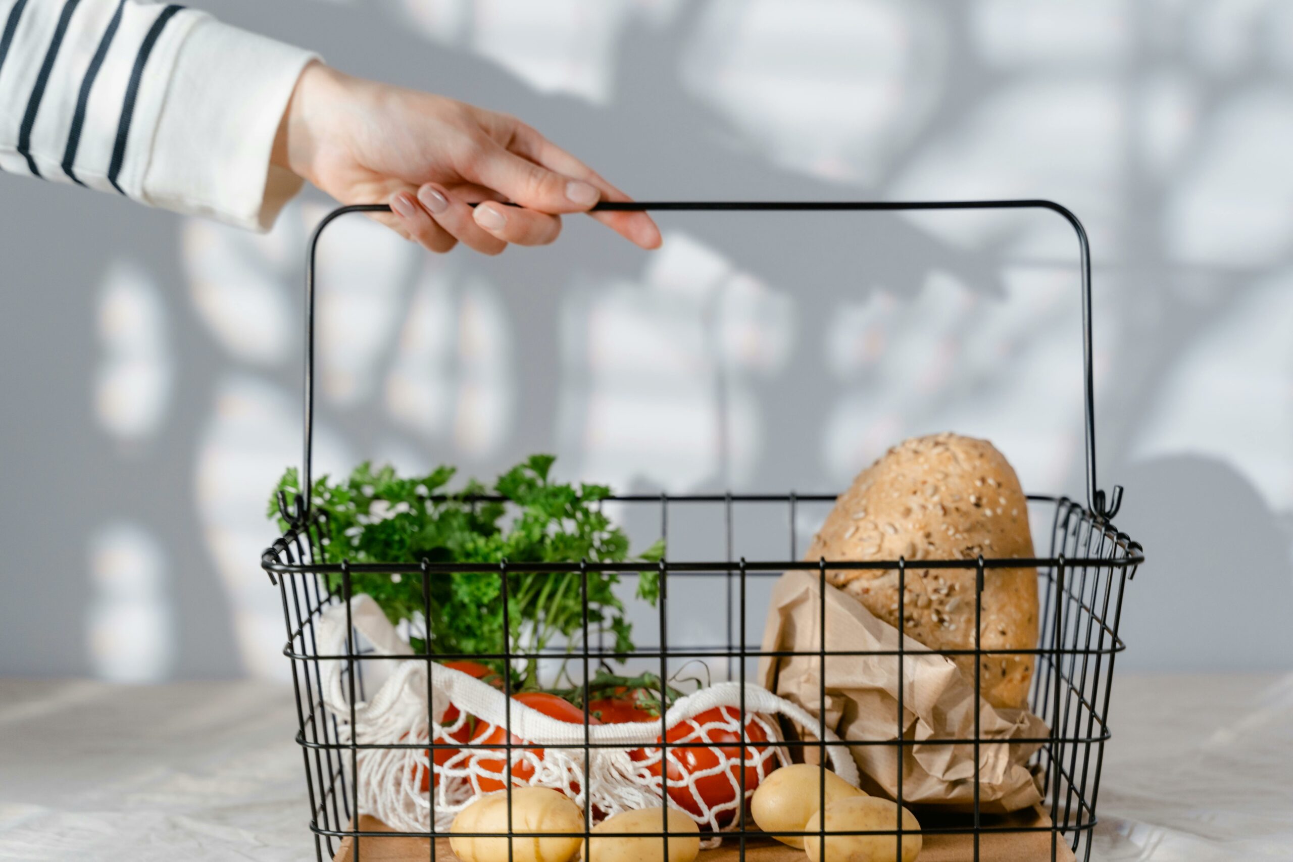 A hand holding a metal basket filled with eco-friendly groceries like bread, vegetables, and parsley.